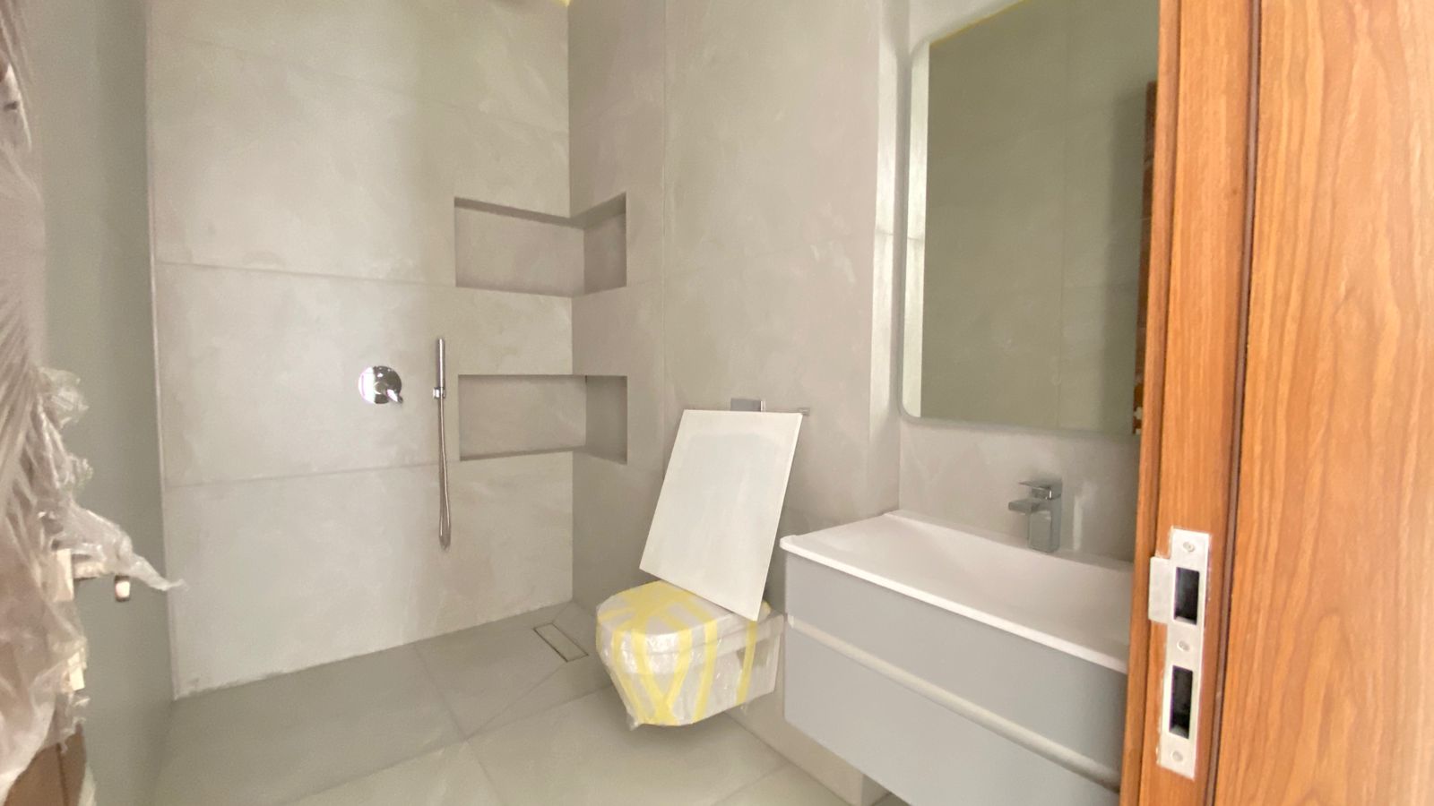 139da8af-67e9-4da3-9cb7-bb65976b4df7 Modern bathroom under construction: large tiled shower area with built-in niches, handheld shower, and a wrapped stool/seat beside a floating vanity and sink mirror.