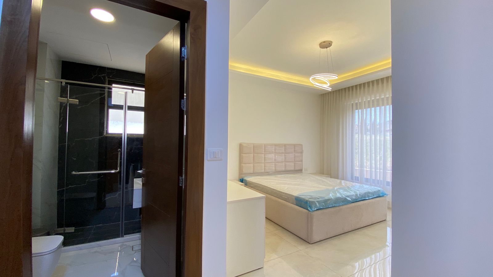 150bd0e0-44ec-44e9-9641-9d63cd0726d5 Bright modern bedroom with a large upholstered bed, sheer curtains, and a circular ceiling light; view into an ensuite bathroom with a glass shower door.