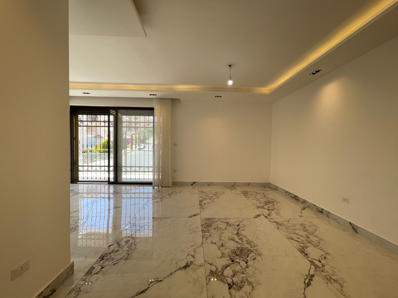 162356c8-ccf8-4f9a-b64d-bd31915bfa7d Empty modern living room with white walls, marble floor, and a sliding glass door to a balcony; warm ceiling lighting.