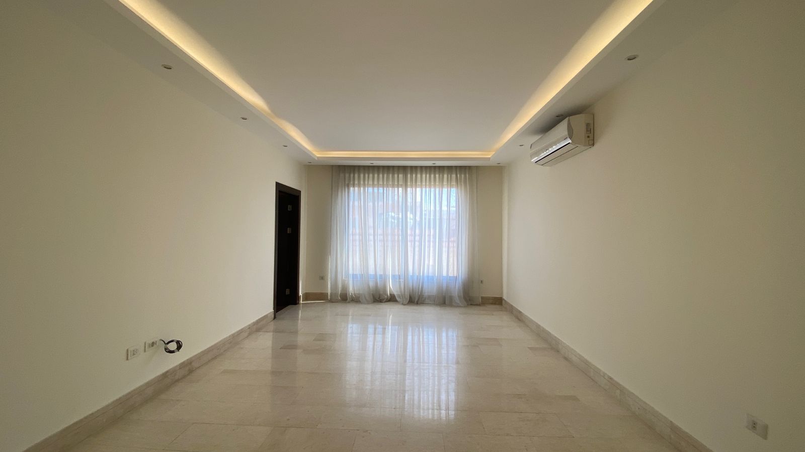 1632b0e7-70ad-482b-bfd2-bb4e87b8e371 Bright empty living room with marble floor, sheer-curtained window at the far end, recessed ceiling lighting, and a wall-mounted air conditioner.