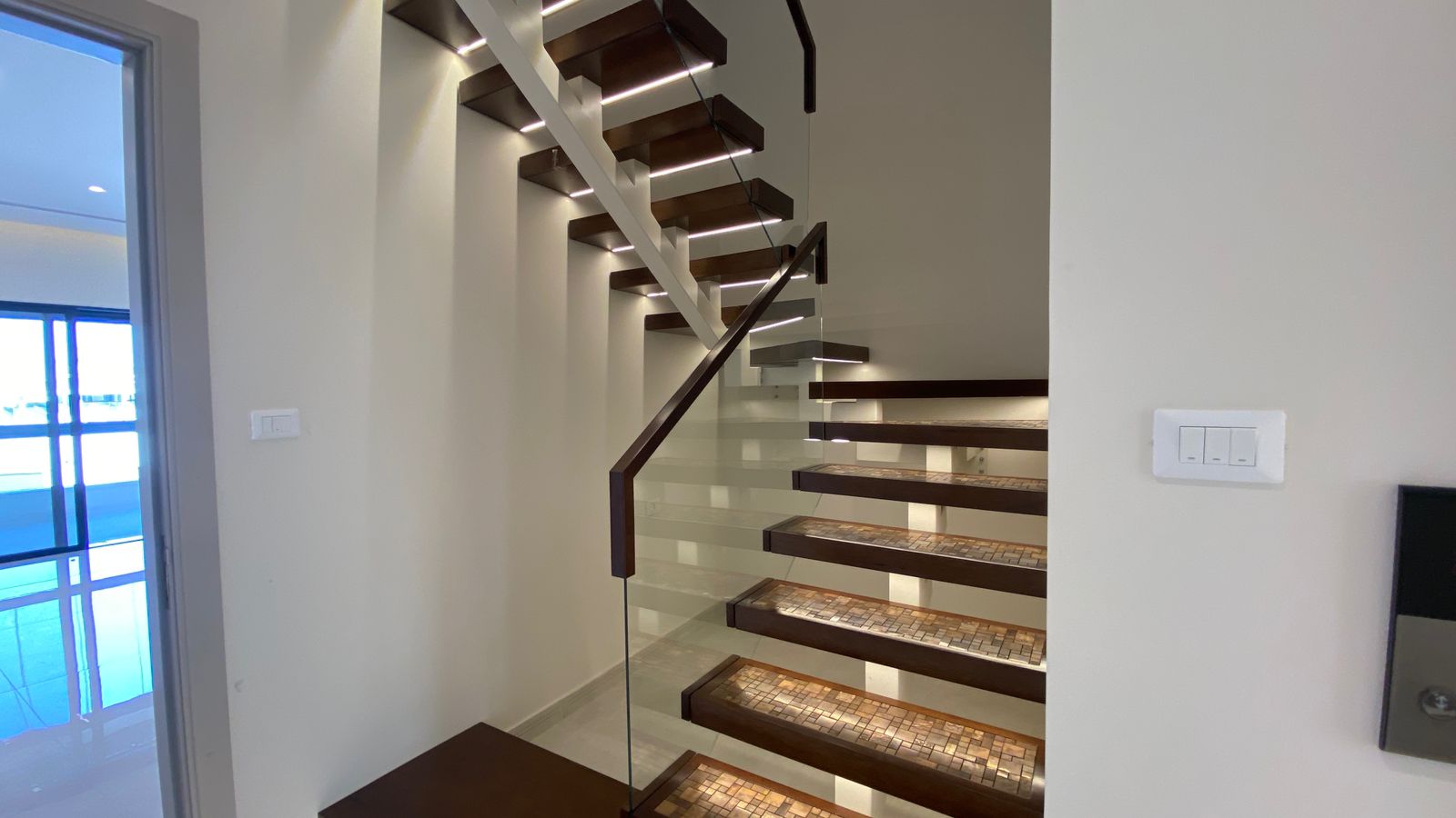 168aa544-df96-4451-a864-954d34942848 Modern floating staircase with dark wood treads and glass balustrade, illuminated by recessed strip lighting.