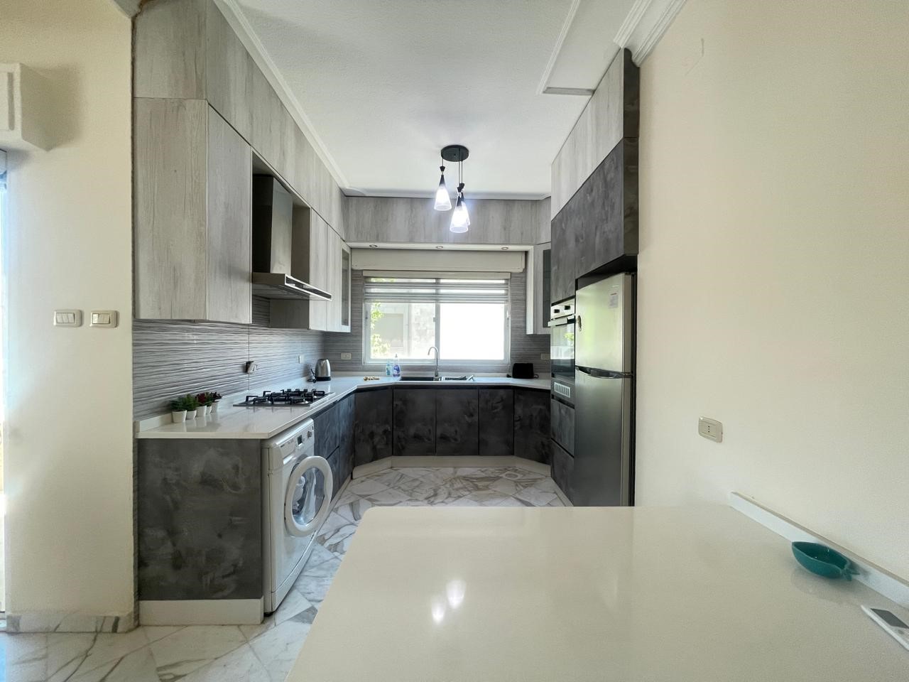 16bba914-080b-46f5-b837-e277ce7988ea L-shaped kitchen with gray lower cabinets, light-toned upper cabinets, white countertops, a washing machine, and a stainless steel refrigerator under a tall cabinet.