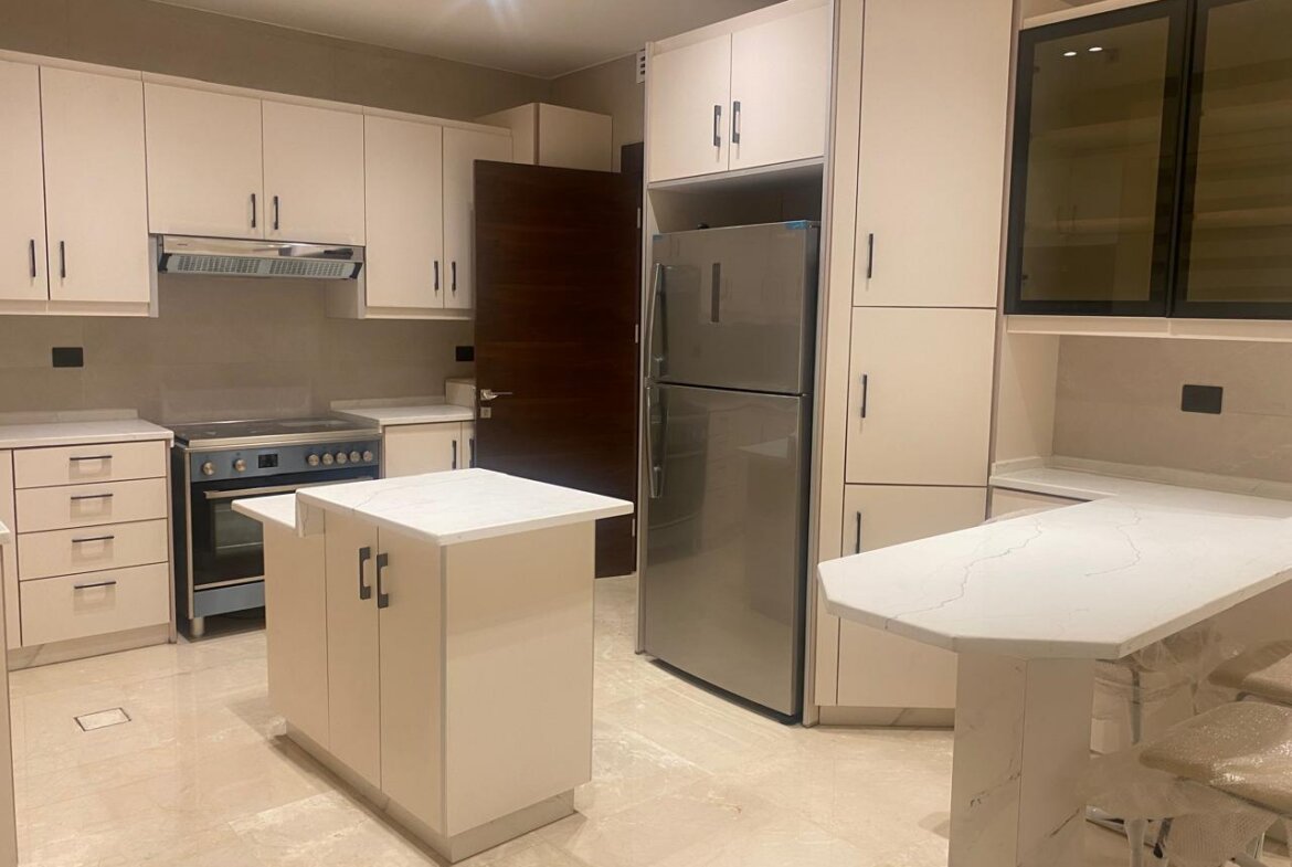 Modern white kitchen with an island, stainless steel refrigerator, and row of upper cabinets; light beige countertops and a glass-front wall cabinet nearby.