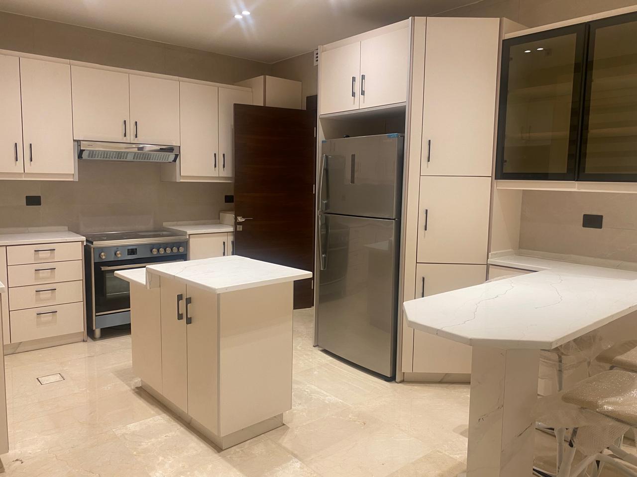 Modern white kitchen with an island, stainless steel refrigerator, and row of upper cabinets; light beige countertops and a glass-front wall cabinet nearby.