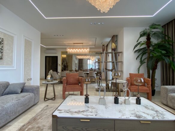 Spacious modern living room with gray sofas, orange armchairs, a marble coffee table, and a tall palm near the curtains; dining area visible in the back.