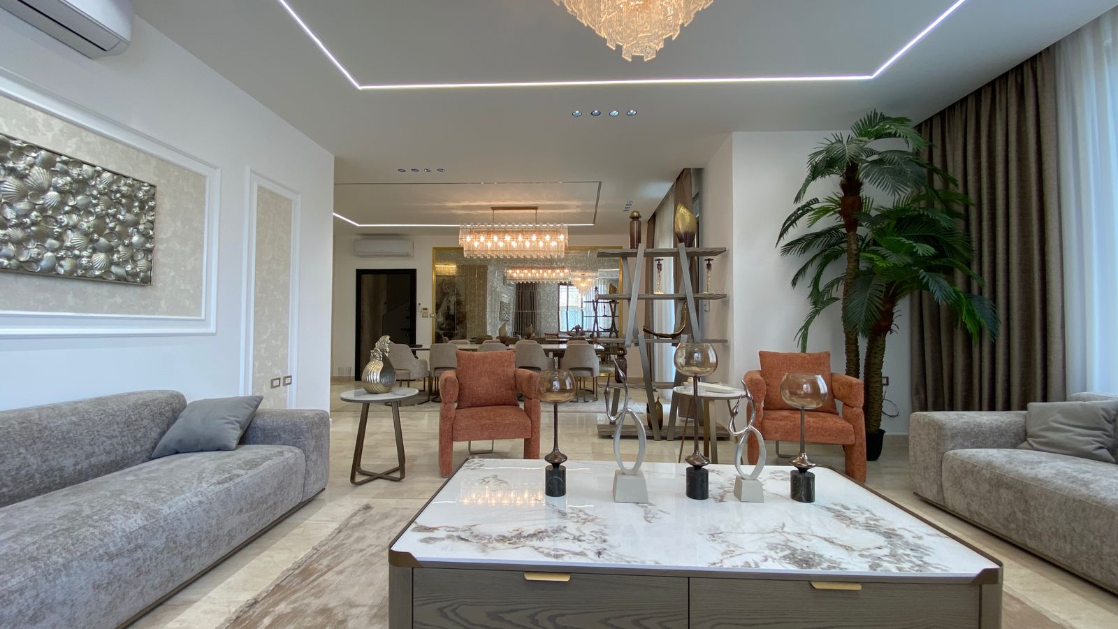 17bf478d-8487-4241-8bcd-bce62dfc9e39 Spacious modern living room with gray sofas, orange armchairs, a marble coffee table, and a tall palm near the curtains; dining area visible in the back.