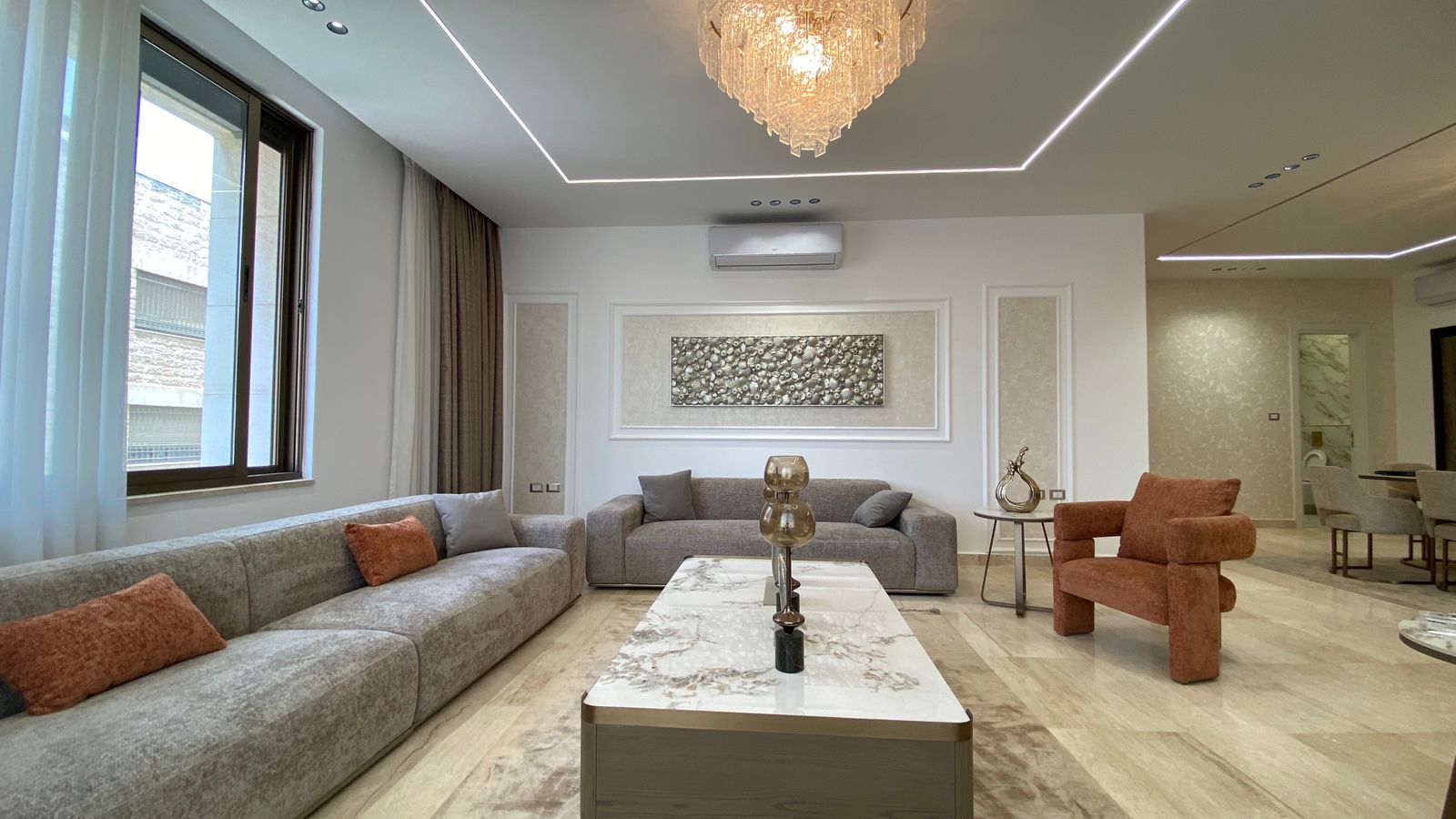 1816f0a7-6b2e-45f5-9cb9-3c3e1ca50c14 Luxurious living room with a gray sectional, rust-orange pillows, a marble coffee table, and a crystal chandelier.