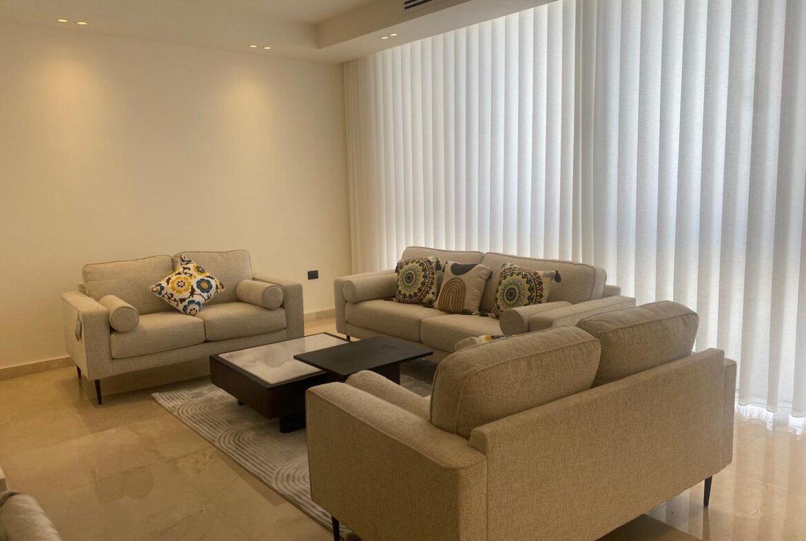 Modern beige living room with three sofas around a dark wood coffee table and colorful patterned cushions under bright vertical blinds.