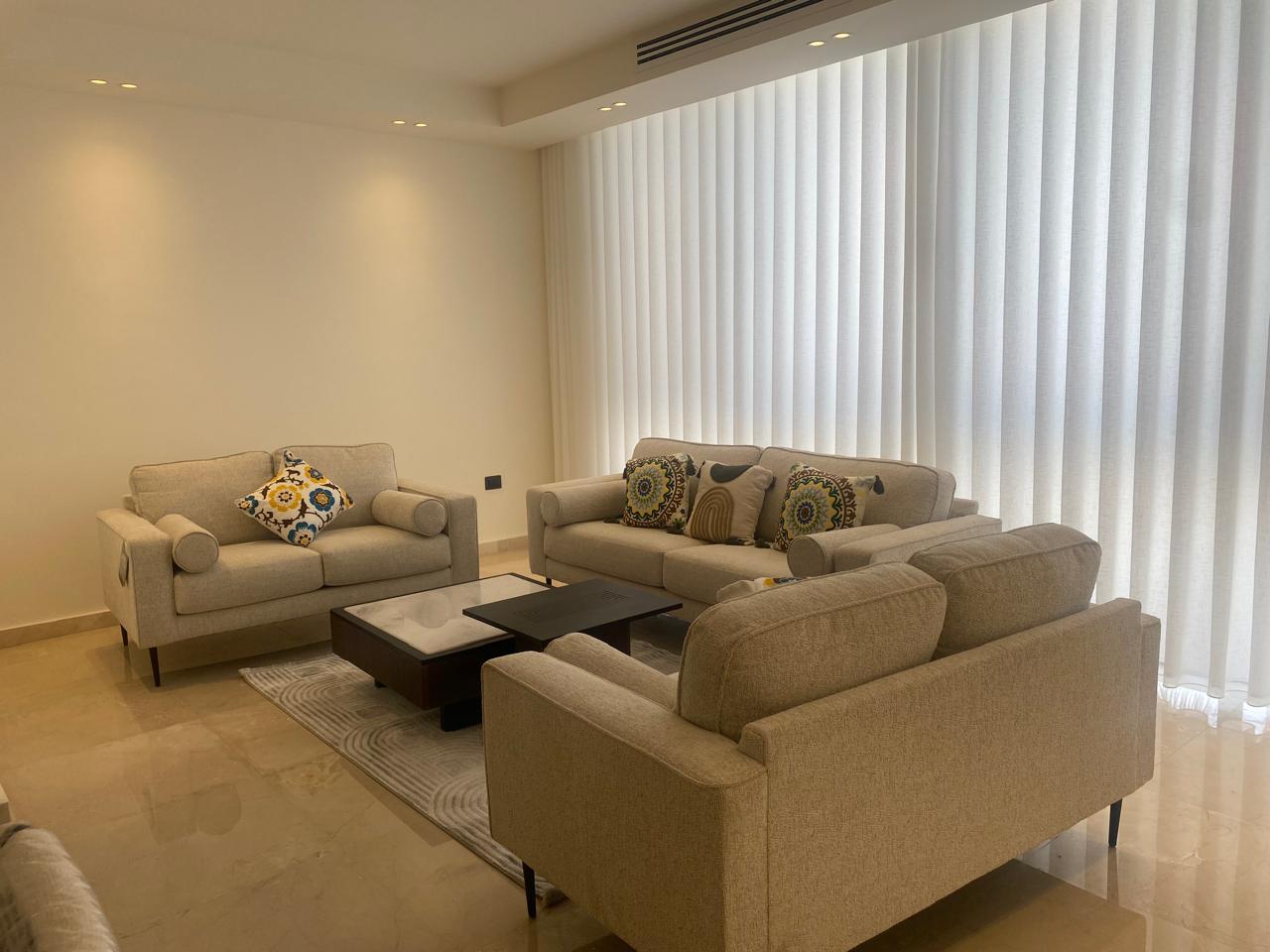Modern beige living room with three sofas around a dark wood coffee table and colorful patterned cushions under bright vertical blinds.