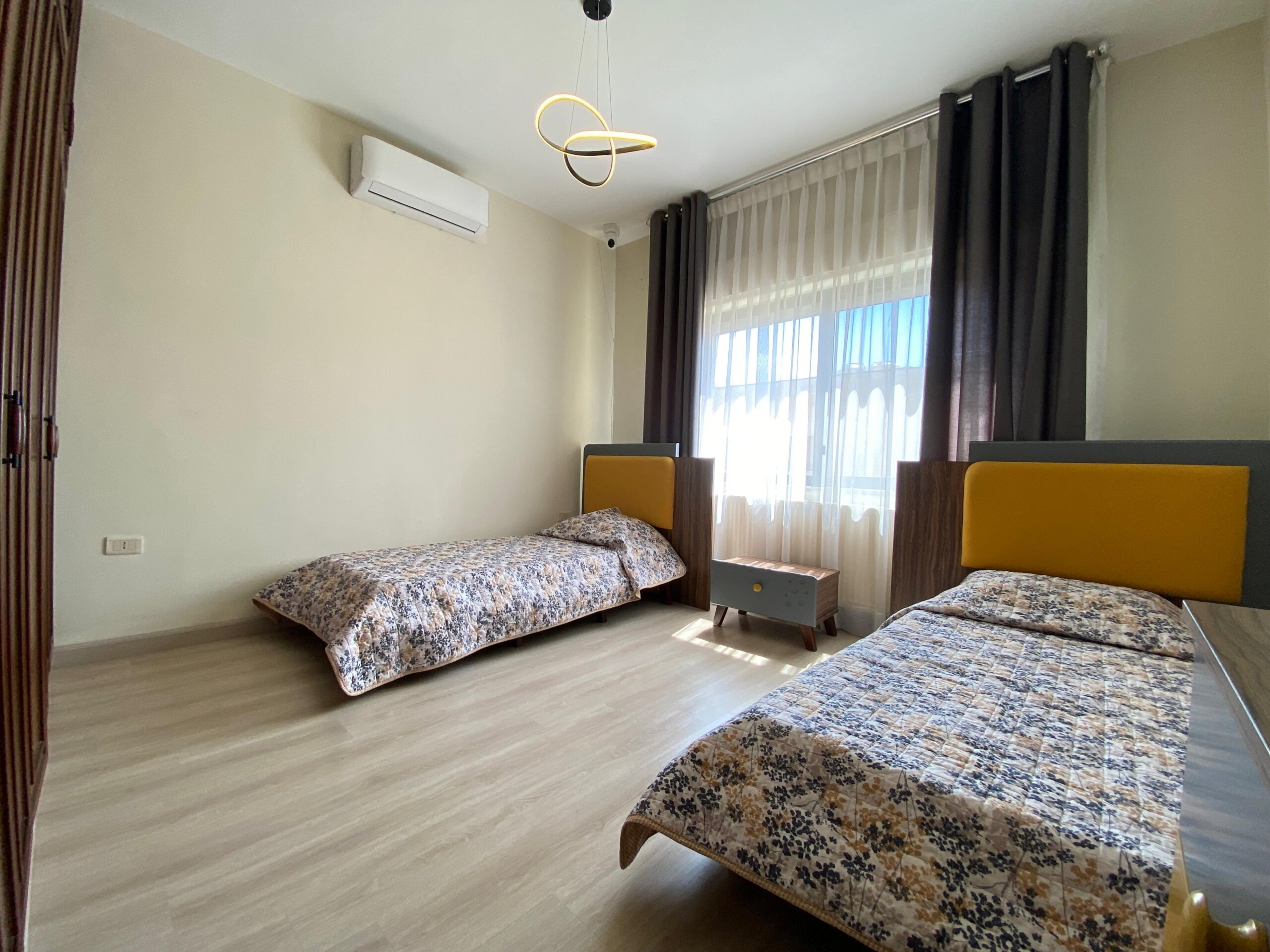 19174871-d765-4379-a42b-c4576cdc33ba Bright bedroom with two twin beds, floral bedding, and mustard-yellow headboards by a large window with sheer curtains; a modern pendant light hangs from the ceiling.