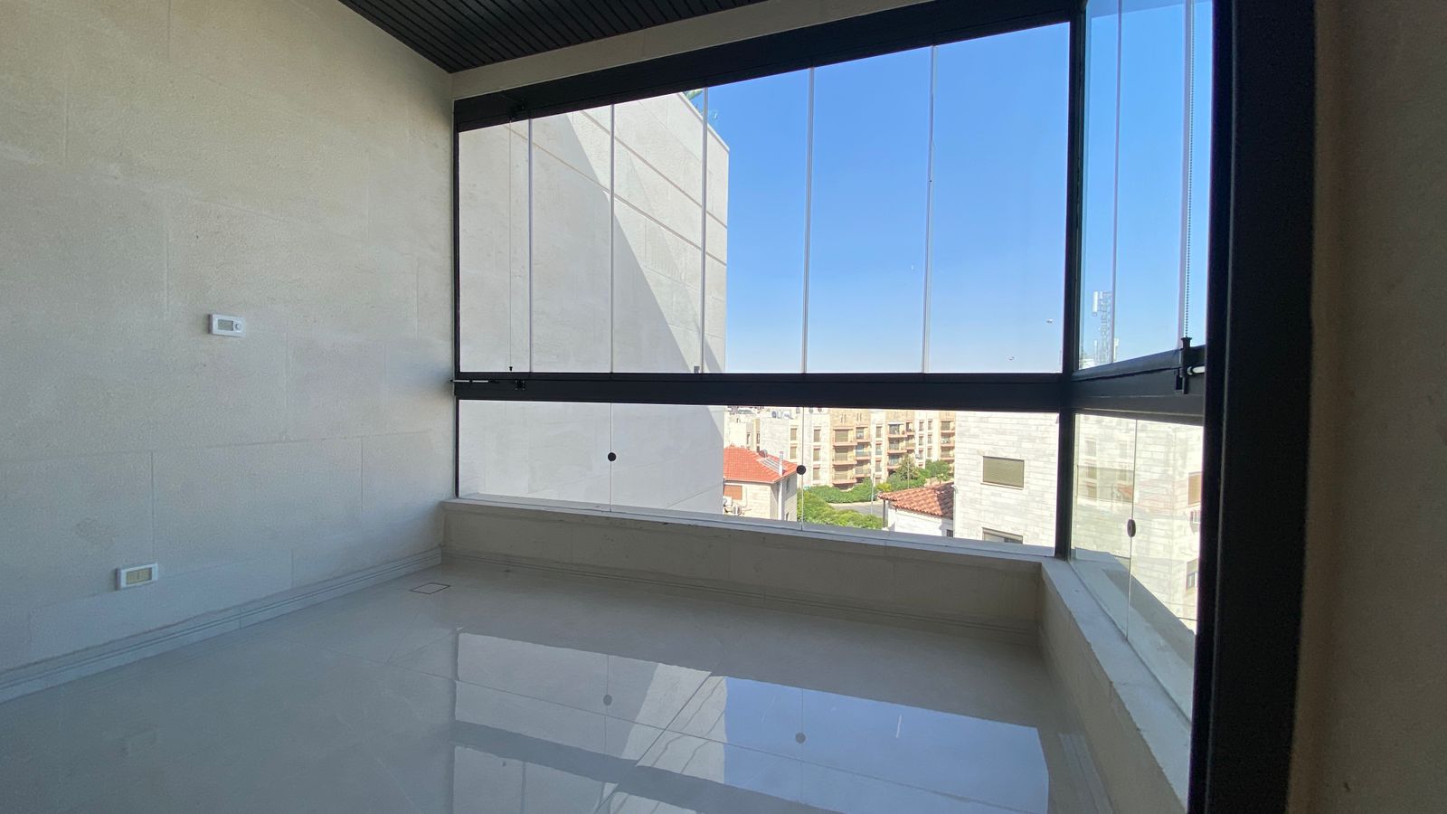 1936a186-24d0-4722-9ee1-b3eae2e1000a Bright, empty balcony or sunroom with large floor-to-ceiling glass windows and a glossy tiled floor, overlooking a cityscape under a clear blue sky.