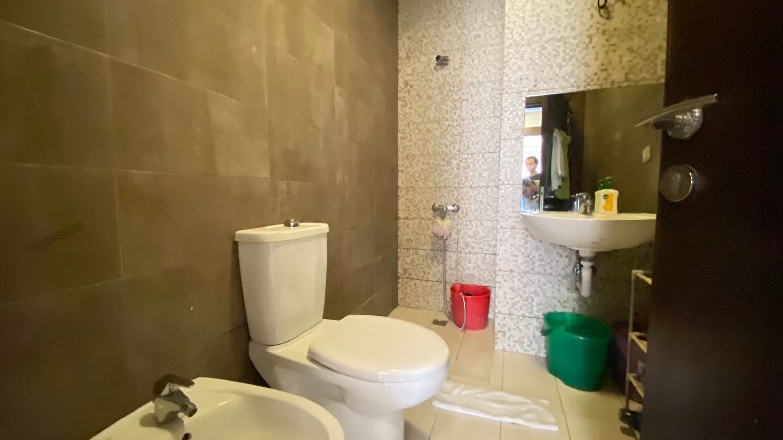 1948830a-37c1-4e7c-bb4d-cf76bfa169d1 Small bathroom with brown tiled walls, white toilet and bidet, and a sink with a large mirror; red and green buckets on the floor and a towel laid out nearby.