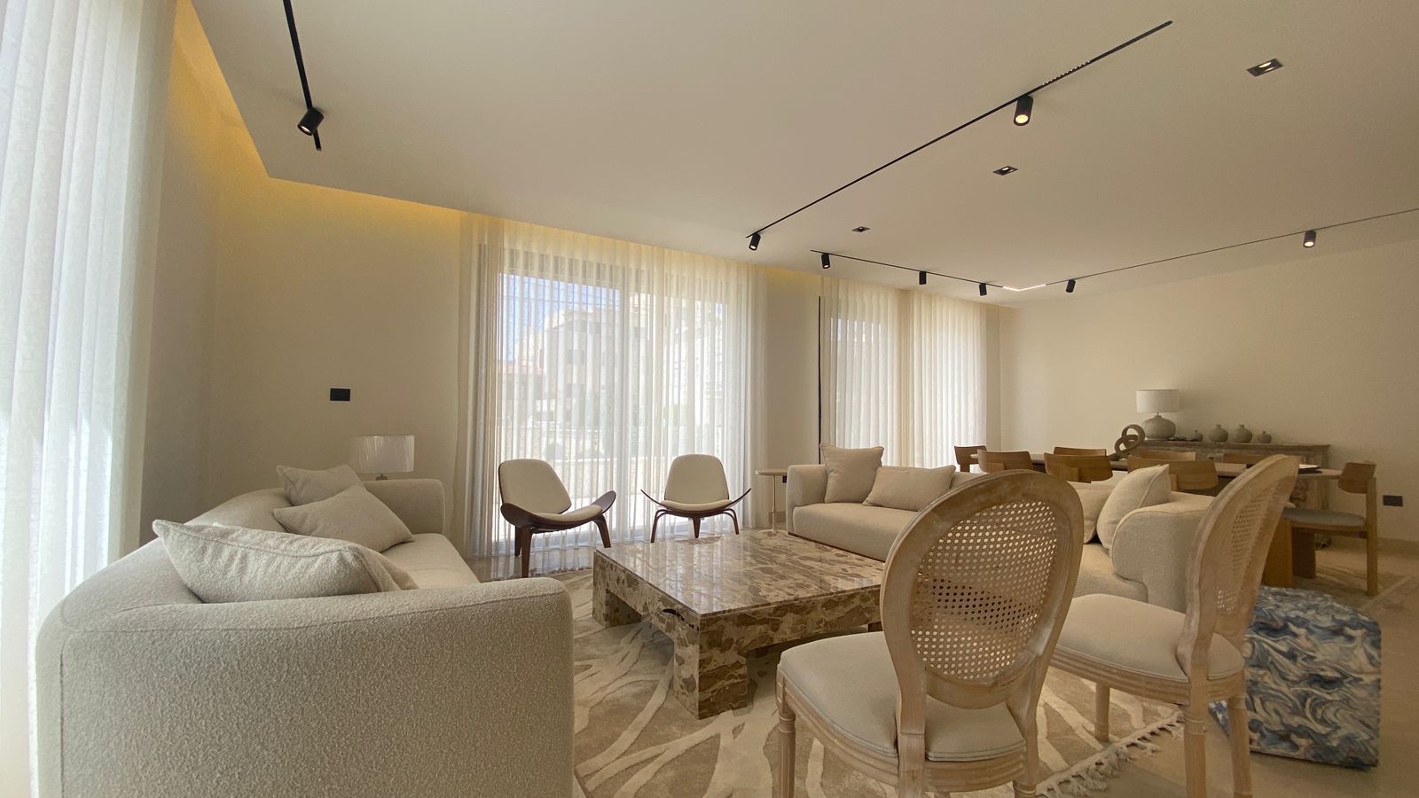 19b4f3e9-b554-4686-91a7-3c340a6db191 Bright, open living room with beige sofas, two white lounge chairs, and a large marble coffee table under track lighting. Substantial sheer-curtain windows let in natural light, with a dining area in the background.