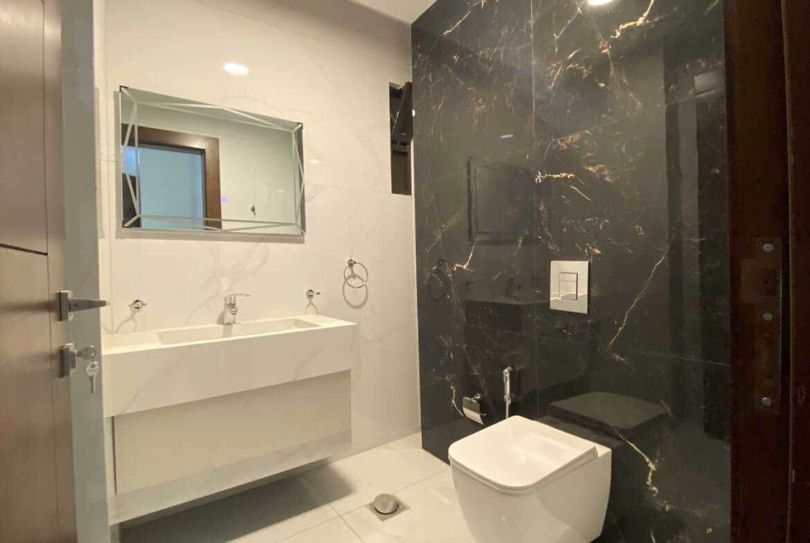 Modern bathroom with a white double-sink vanity, rectangular mirror, and a black marble wall behind the toilet area.