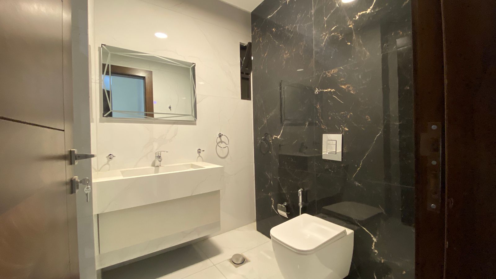 19ba20ff-62b8-479b-b748-59cb7b3a711e Modern bathroom with a white double-sink vanity, rectangular mirror, and a black marble wall behind the toilet area.
