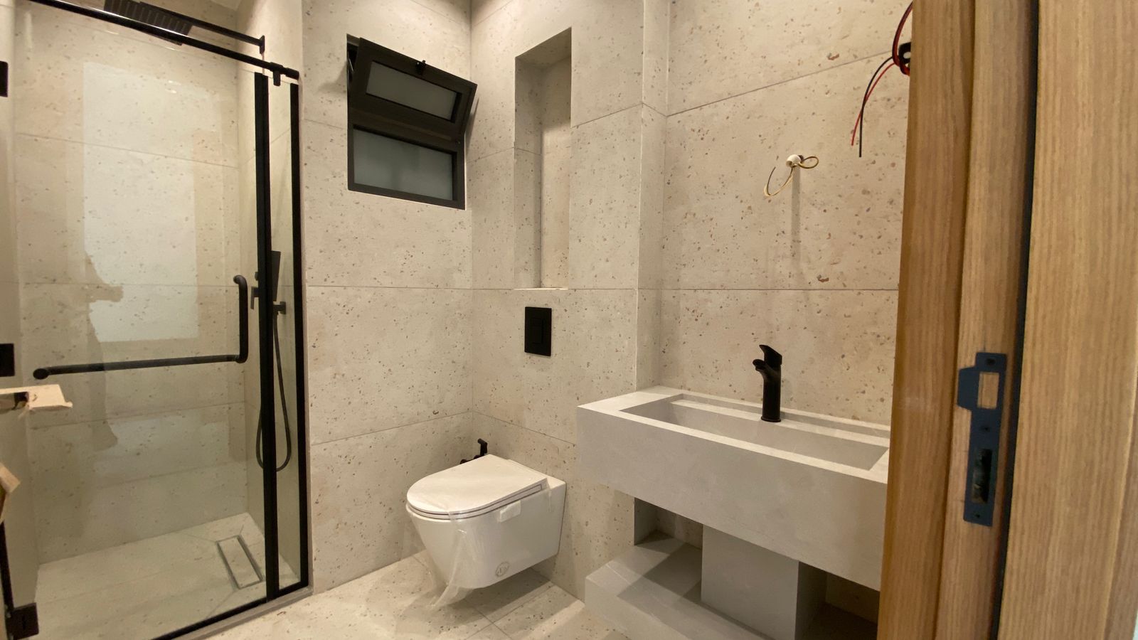 19f82b01-ef53-46b7-b2f2-a42909957adf Contemporary bathroom under construction with a glass-framed shower, wall-mounted toilet, and a concrete sink; wiring exposed near the doorway.