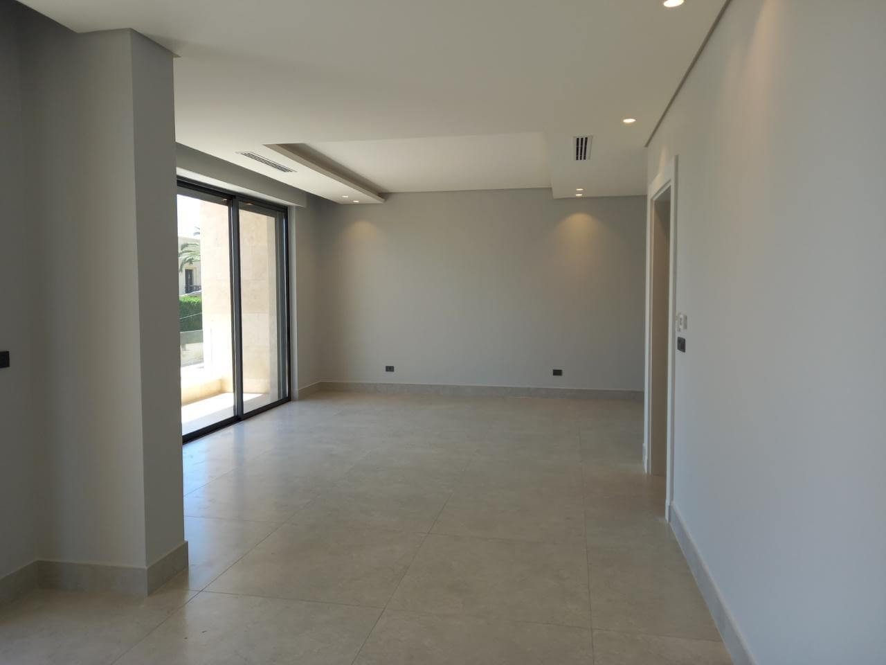 19fbc04b-d69f-4460-8f22-52c8e983cdba Empty modern living room with beige tiled floor, light gray walls, and large sliding glass doors to a patio/outdoor space at left edge of the room.