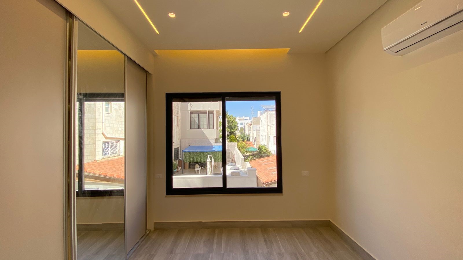 1b43a72f-f20c-49aa-a1c1-11072f93ae6e Empty modern bedroom with a large central window, mirrored sliding closet doors on the left, light wood flooring, and a wall-mounted AC unit on the right.