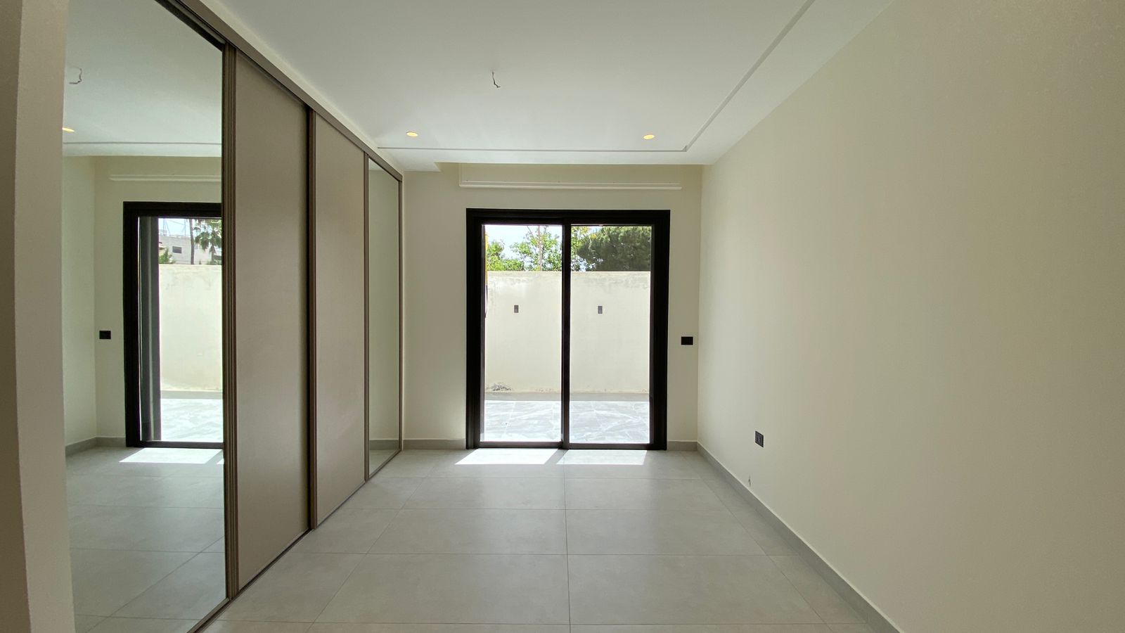 1bcfa824-73b4-4ed3-9da6-eda23f699e22 Empty modern bedroom with mirrored sliding doors on the left and a glass door opening to a courtyard outside.