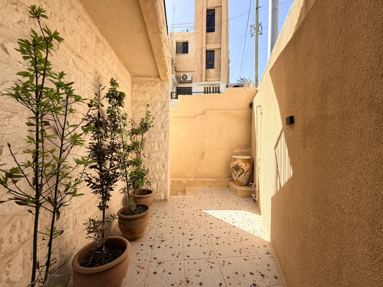 1bdcabff-cecb-4604-8374-11011d518660 Sunlit beige courtyard with tall potted trees along the left wall, patterned tile floor, and a large decorative ceramic vase near the steps at the far end.