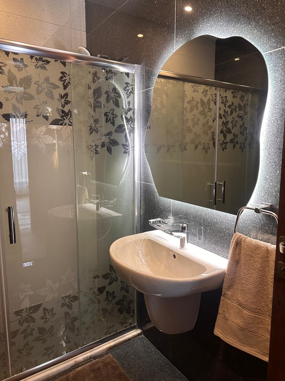 1c161195-e30c-4e85-b6a8-2bfd08a6f06a Modern bathroom with frosted glass shower doors featuring a botanical leaf pattern, and a wall-mounted sink with a backlit mirror.