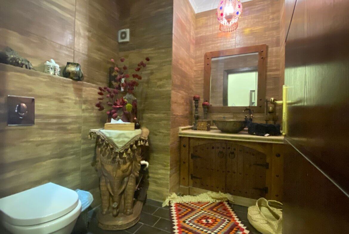 1d428970-99a9-4117-bdde-a24b86b03d2d Rustic bathroom with wooden paneled walls, a carved elephant pedestal, tissue box, and a wooden vanity with sink and mirror. A colorful rug sits on the floor.