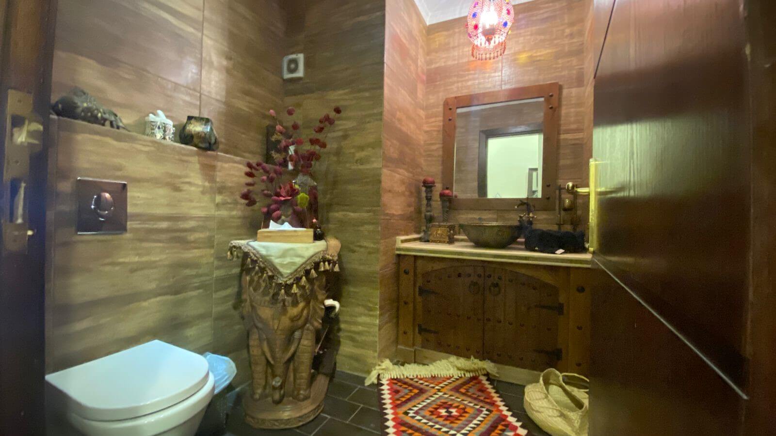 1d428970-99a9-4117-bdde-a24b86b03d2d Rustic bathroom with wooden paneled walls, a carved elephant pedestal, tissue box, and a wooden vanity with sink and mirror. A colorful rug sits on the floor.