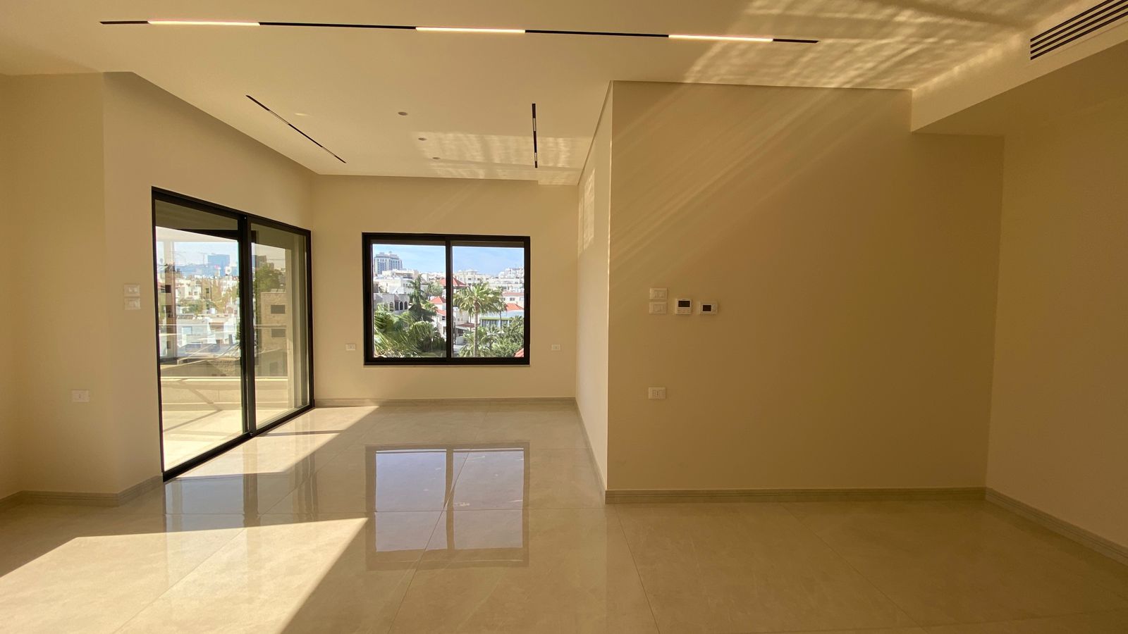 1d8cf3ff-3436-49c5-9fb7-41be334ceb84 Bright empty living room with large sliding glass doors to a balcony, beige walls, and glossy tiled floor reflecting sunlight from outside.