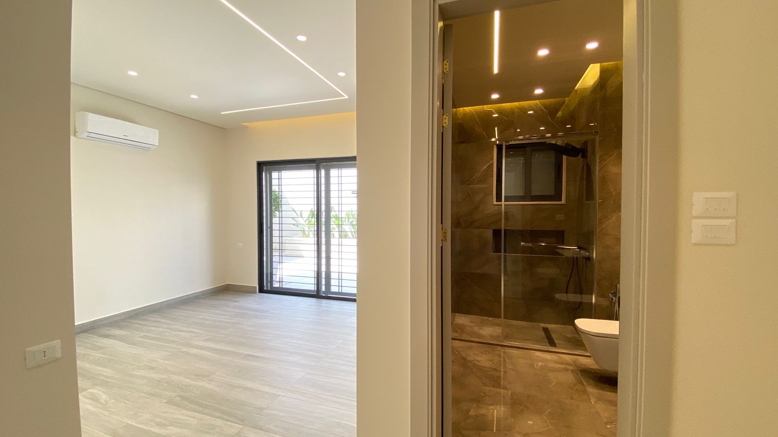 1d9a0476-20e1-4bcd-bc65-35645aa3c338 Modern living area with grey wood floors, white walls, and a wall-mounted air conditioner; sliding doors to the outside.
