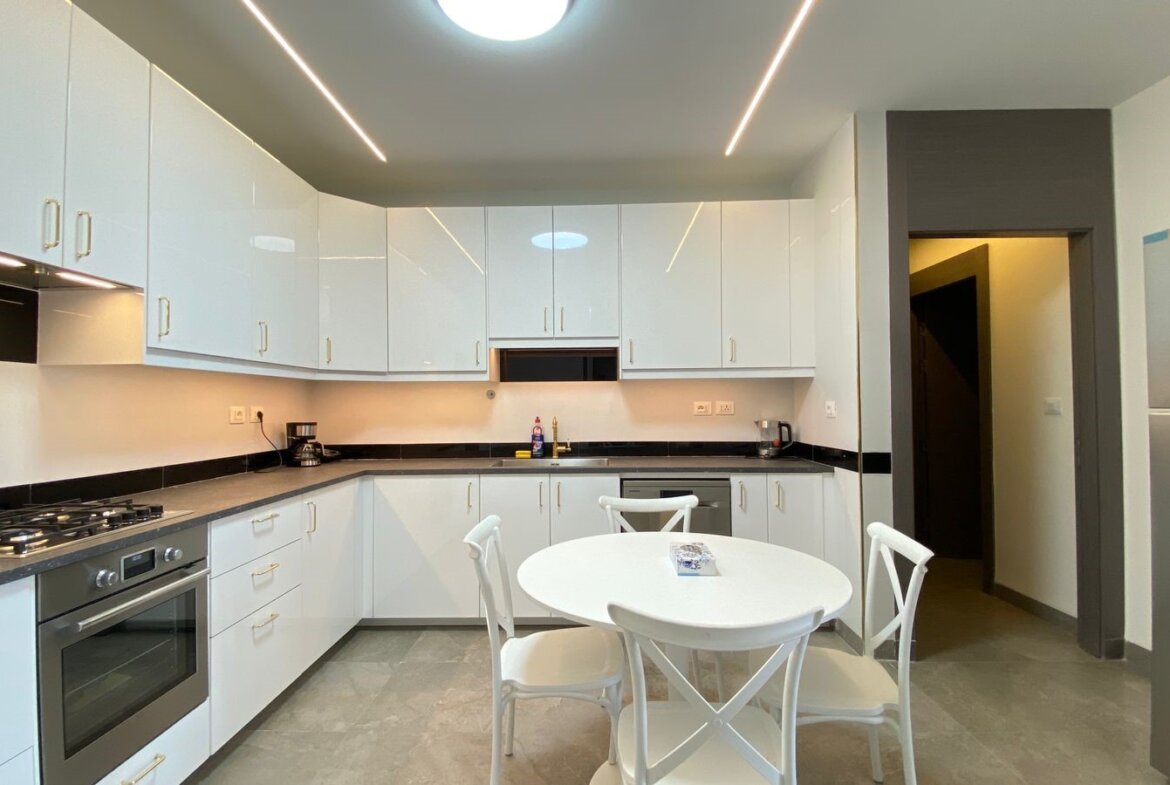 Modern U-shaped kitchen with white glossy cabinets, black countertops, and stainless steel appliances including a built-in oven and gas stove. A white round table with four chairs sits in the center.