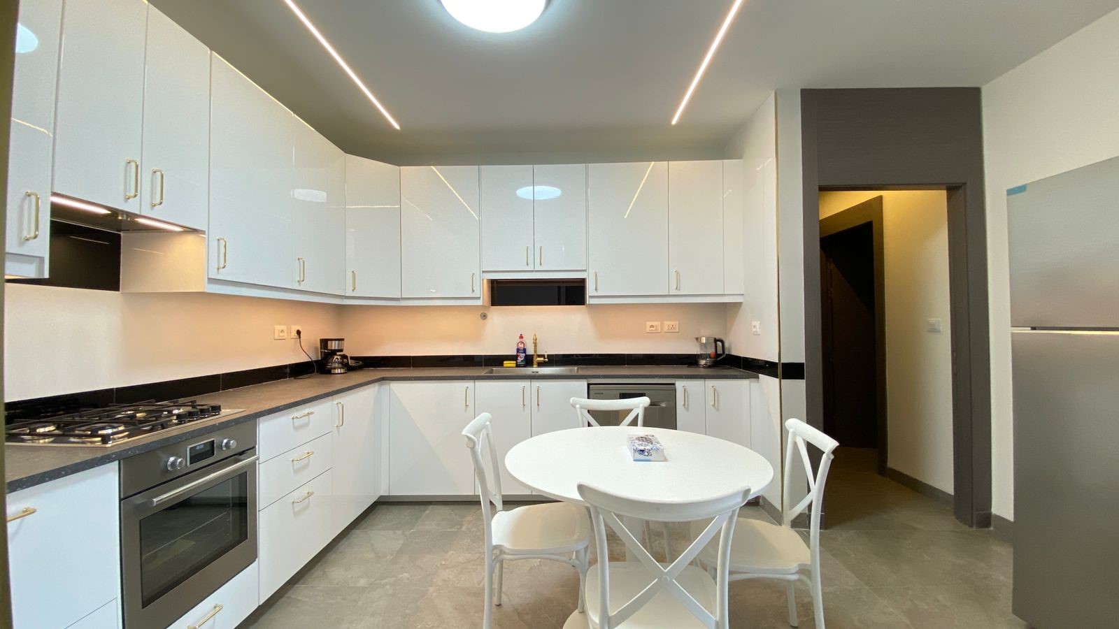 1de6392e-1998-4fa5-92c6-1ae3d46401d1 Modern U-shaped kitchen with white glossy cabinets, black countertops, and stainless steel appliances including a built-in oven and gas stove. A white round table with four chairs sits in the center.