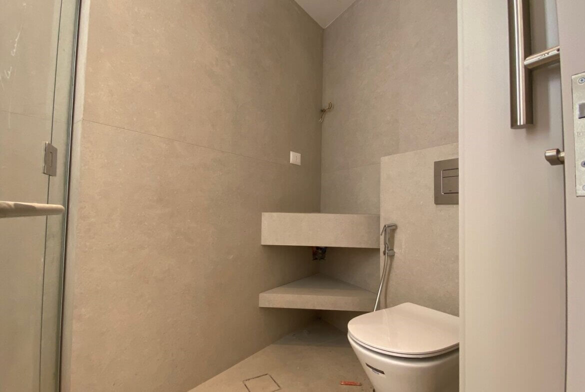 1e157ace-db27-48d6-b8e4-522bfdb3abe2 Minimalist bathroom with concrete walls, a glass shower door on the left, and a wall-mounted toilet on the right, plus two floating shelves and exposed plumbing details in a neutral-toned space