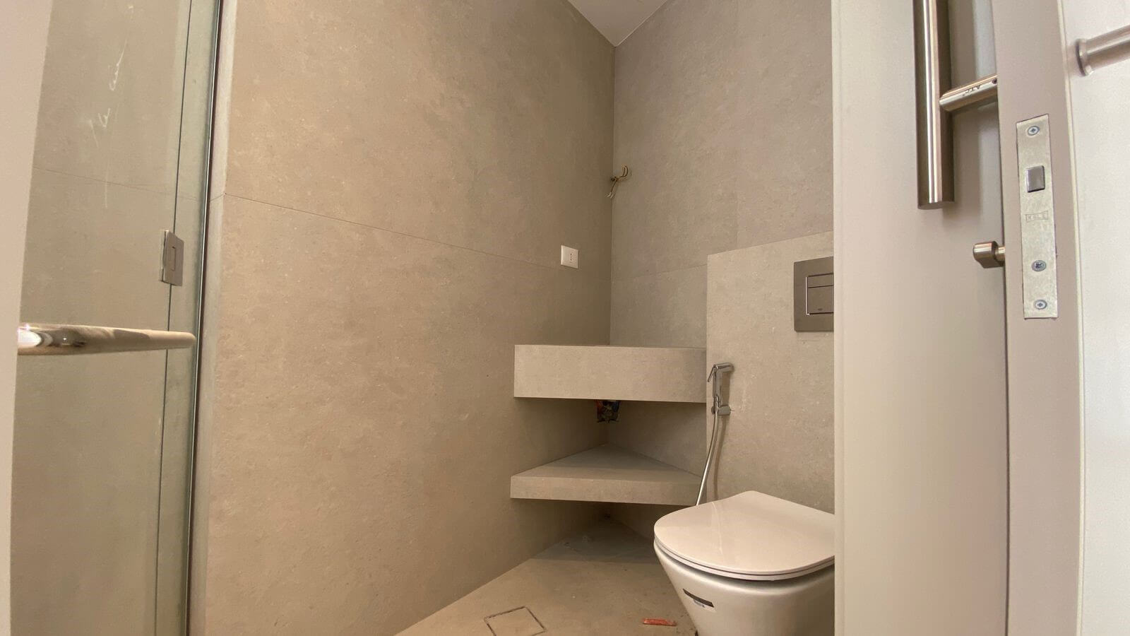 1e157ace-db27-48d6-b8e4-522bfdb3abe2 Minimalist bathroom with concrete walls, a glass shower door on the left, and a wall-mounted toilet on the right, plus two floating shelves and exposed plumbing details in a neutral-toned space