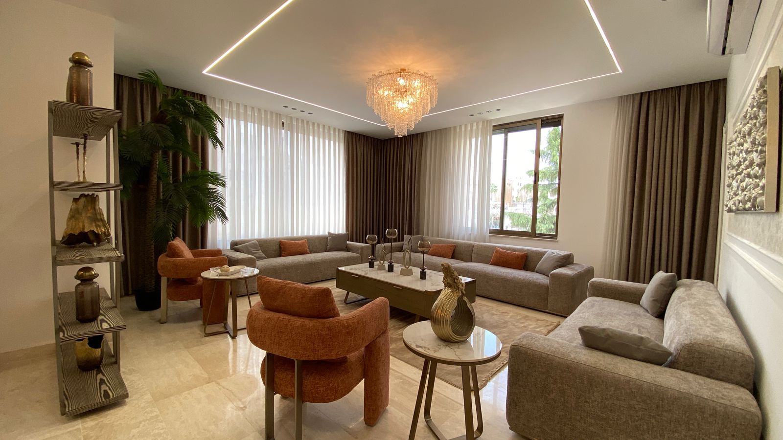 1e704c6c-d8eb-4f53-921a-07b191f611a4 L-shaped gray sofa seating around a marble coffee table in a bright living room with a crystal chandelier.