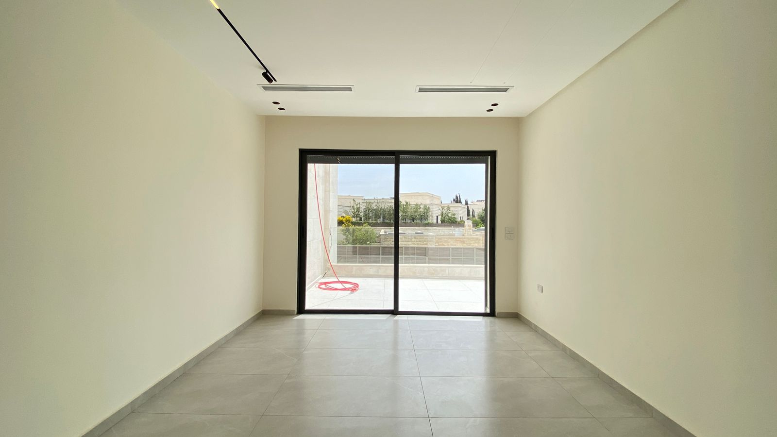 1f2a18cf-4050-4a97-ae38-c99781806ce0 Empty rectangular room with tiled gray floor and sliding glass doors opening to an outdoor patio; red hose visible outside.