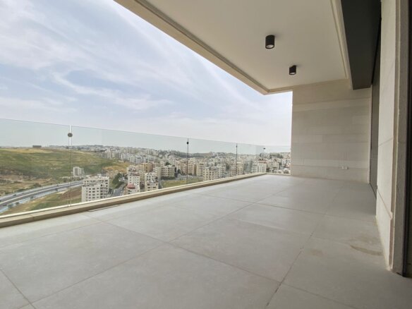 Spacious balcony with glass railing overlooking a city and hills in the distance, under a covered ceiling.