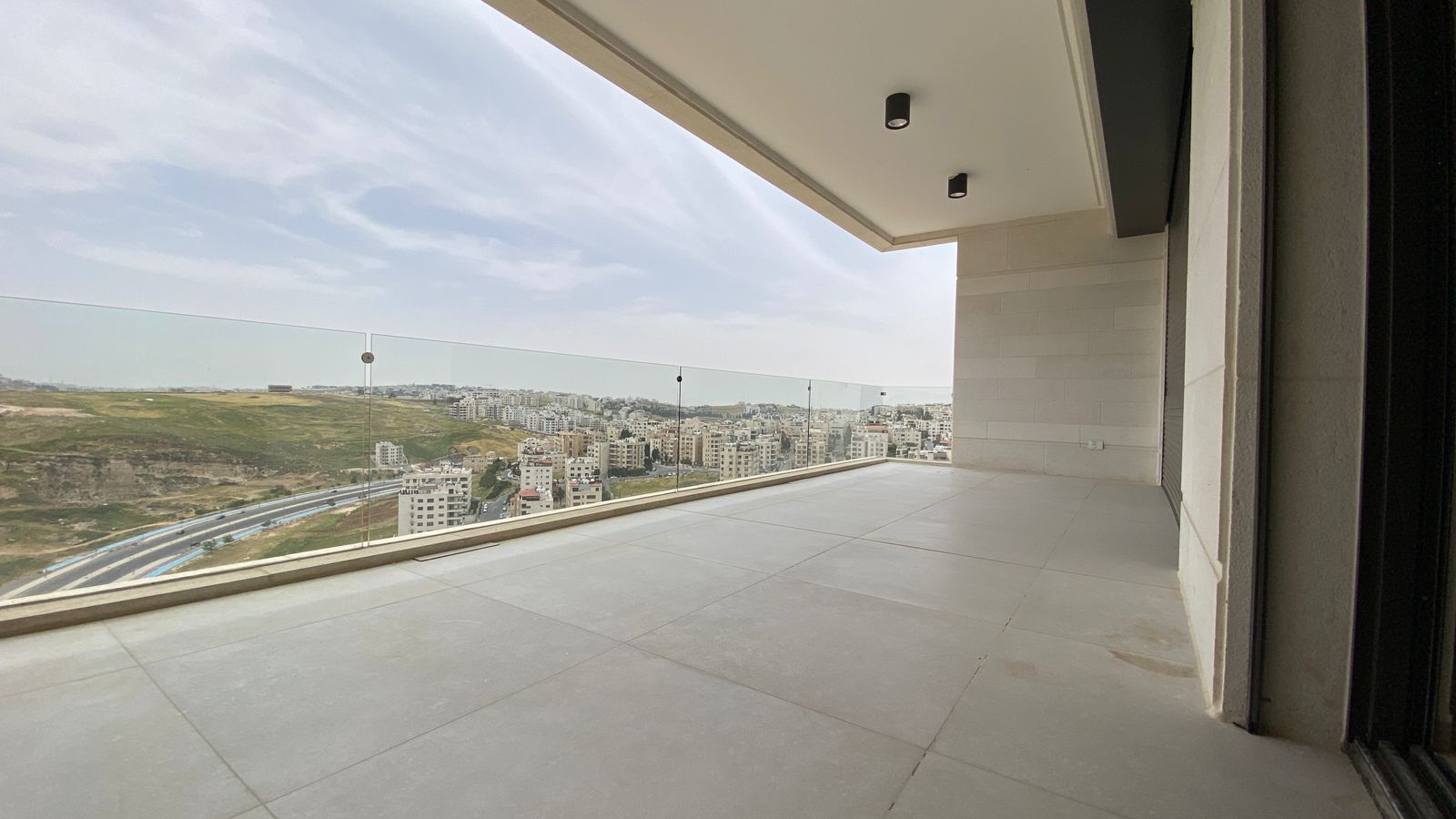 1f622042-6174-4211-a1e0-f7d1daafb038 Spacious balcony with glass railing overlooking a city and hills in the distance, under a covered ceiling.
