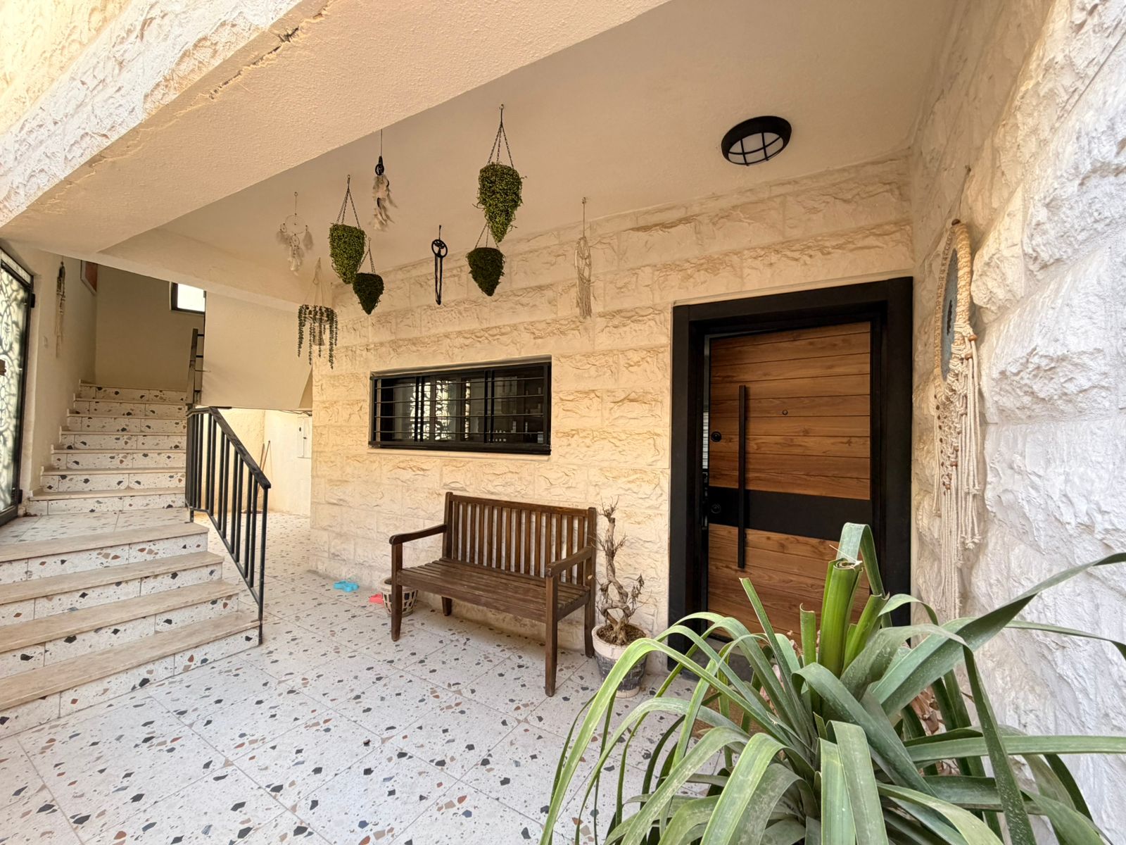 1fadb2df-a215-4db1-bb8b-6a0ef06004c0 Outdoor entryway with white stone walls, wooden front door, bench, stairs on the left, and hanging planters above.