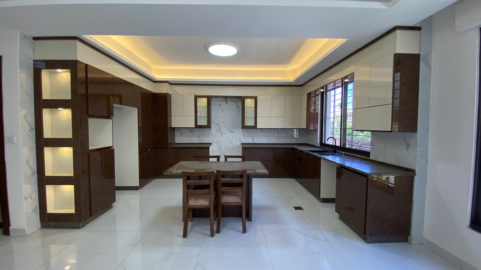 20133e9f-36ba-45d5-8e35-78b452f6831d Modern L-shaped kitchen with brown and cream cabinets, marble backsplash, and a central dining table with four chairs.