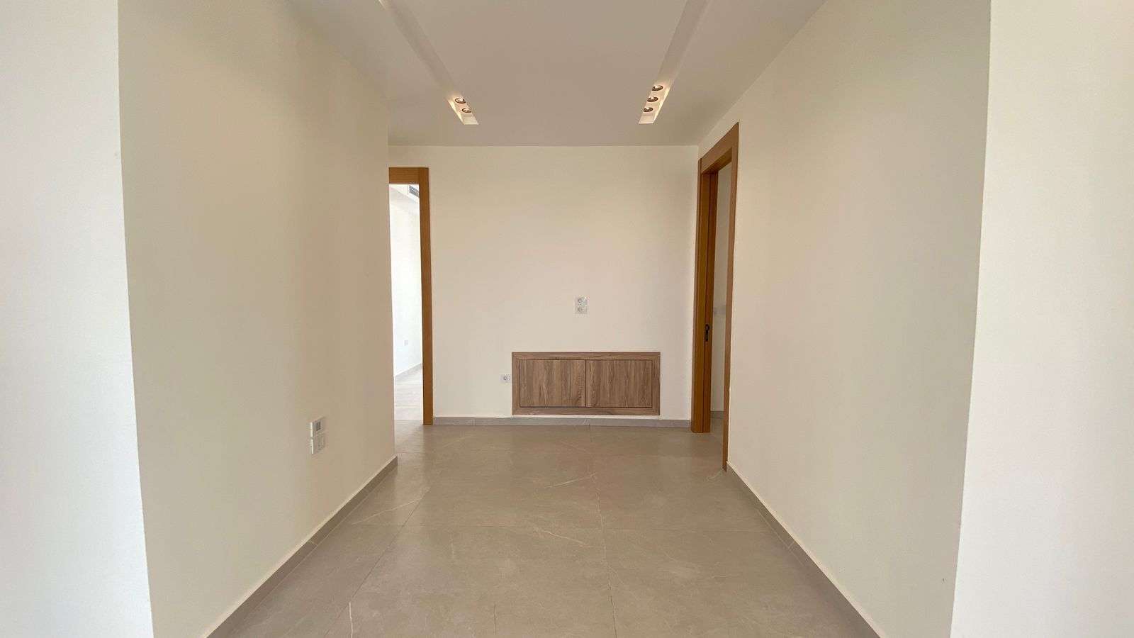 201a3373-adfa-4c00-9483-116c41c4b4be Empty hallway with white walls, gray tiled floor, and wooden door frames; a built-in wood cabinet on the far wall under a switch panel; bright track lighting overhead.