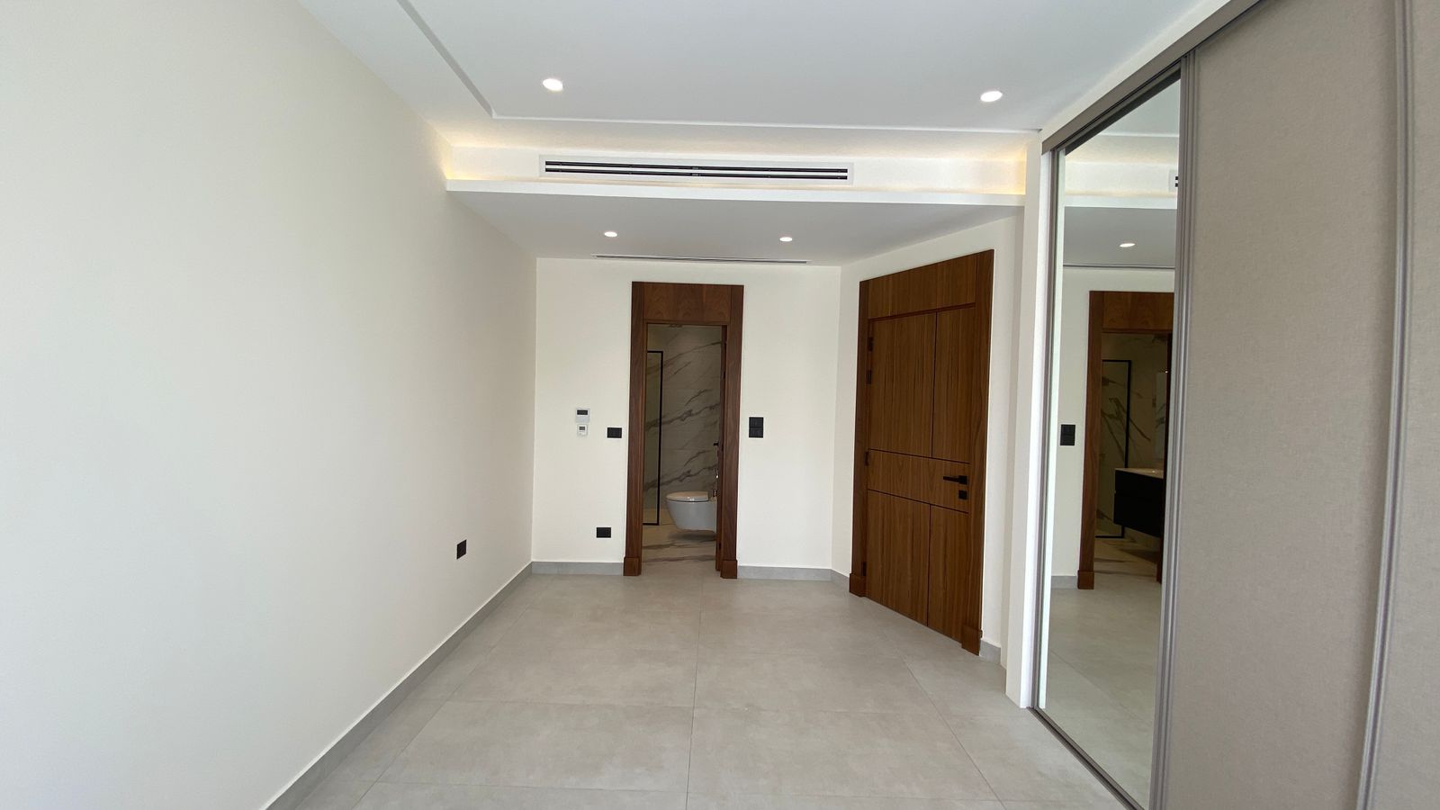 213a493c-2872-4c07-8085-7f507aa77216 Modern hallway with beige walls, tiled floor, wooden doors at the end, and a large mirrored wardrobe on the right; bathroom visible through an open door.