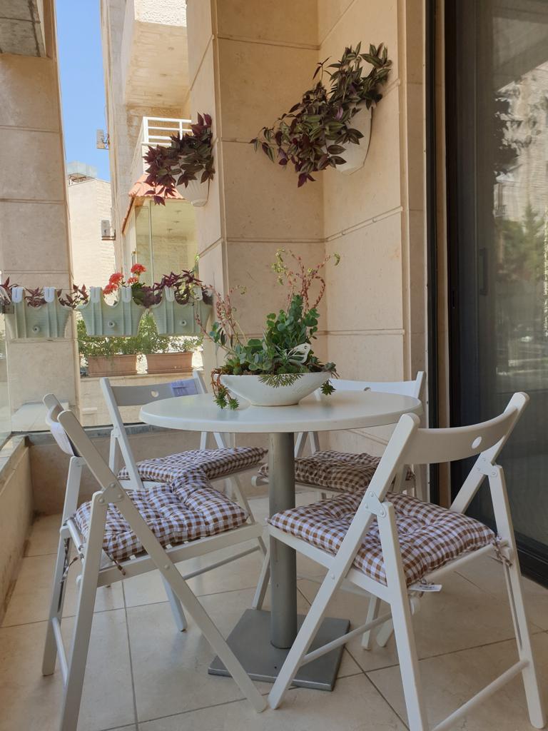 21a6e540-17c6-42ce-b4b9-041bb5c1f1c5 White round patio table with four folding chairs and gingham cushions on a sunlit balcony with hanging and potted plants around it.