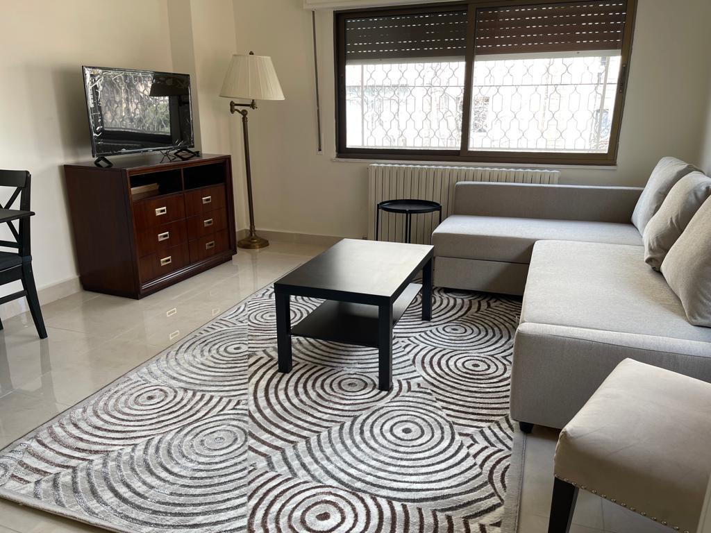 Living room with a gray sectional sofa, black coffee table, and a circular-pattern rug; a TV sits on a dark wood cabinet beside a tall floor lamp. Window with metal grate and a radiator are visible in the background.