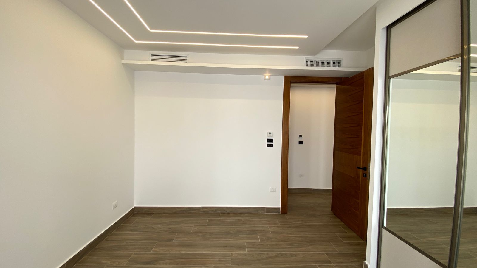 21af2d5c-73e5-4d76-9a56-6f55b7b047e0 Empty modern room with wood-toned tile flooring, white walls, and a wooden door slightly ajar.