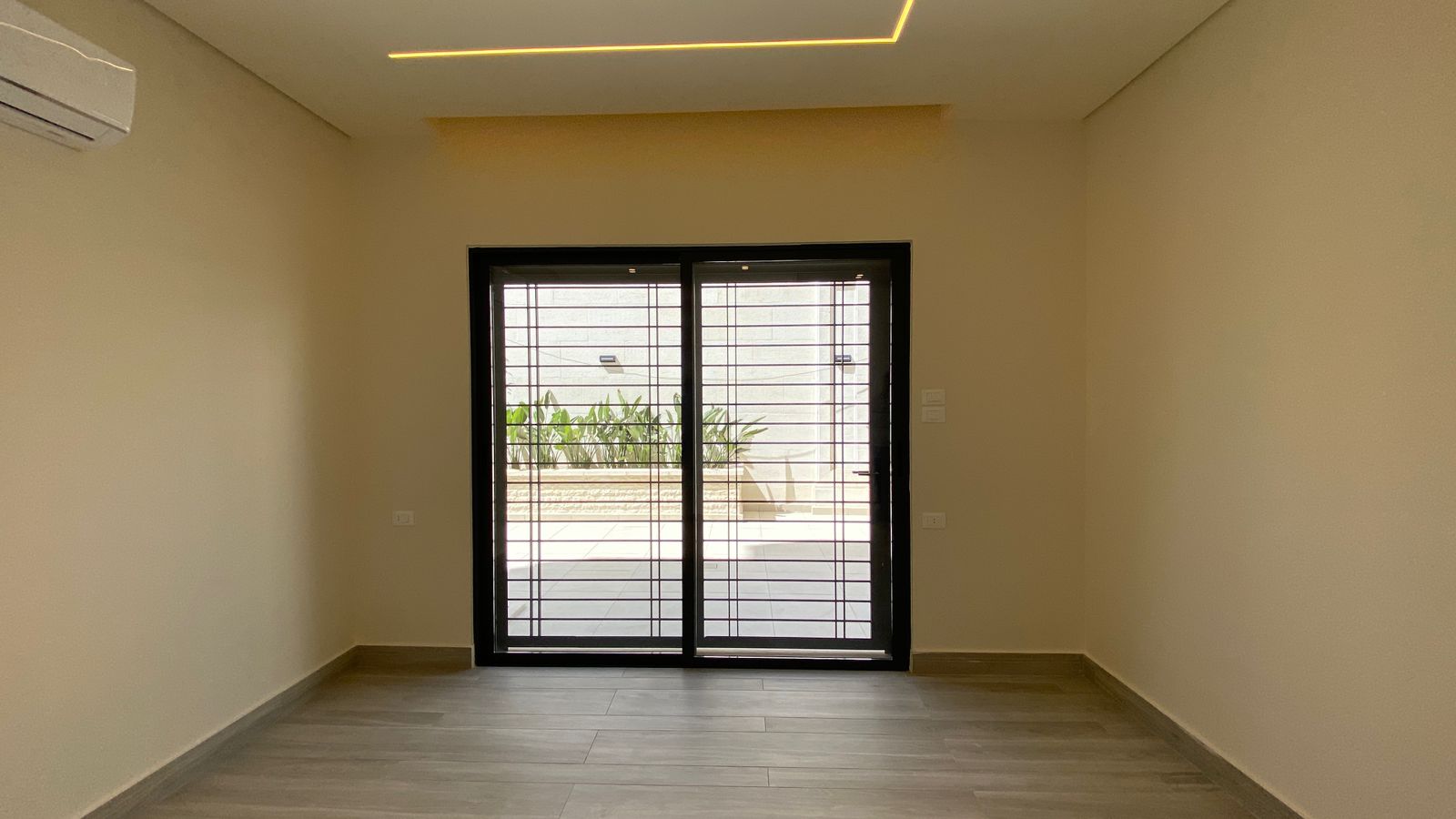 232c26f5-4a61-4a2f-9dc2-e012c6a3da40 Empty modern room with light wood flooring and beige walls, featuring a black-framed sliding glass door with metal bars to a sunny outdoor patio with plants.