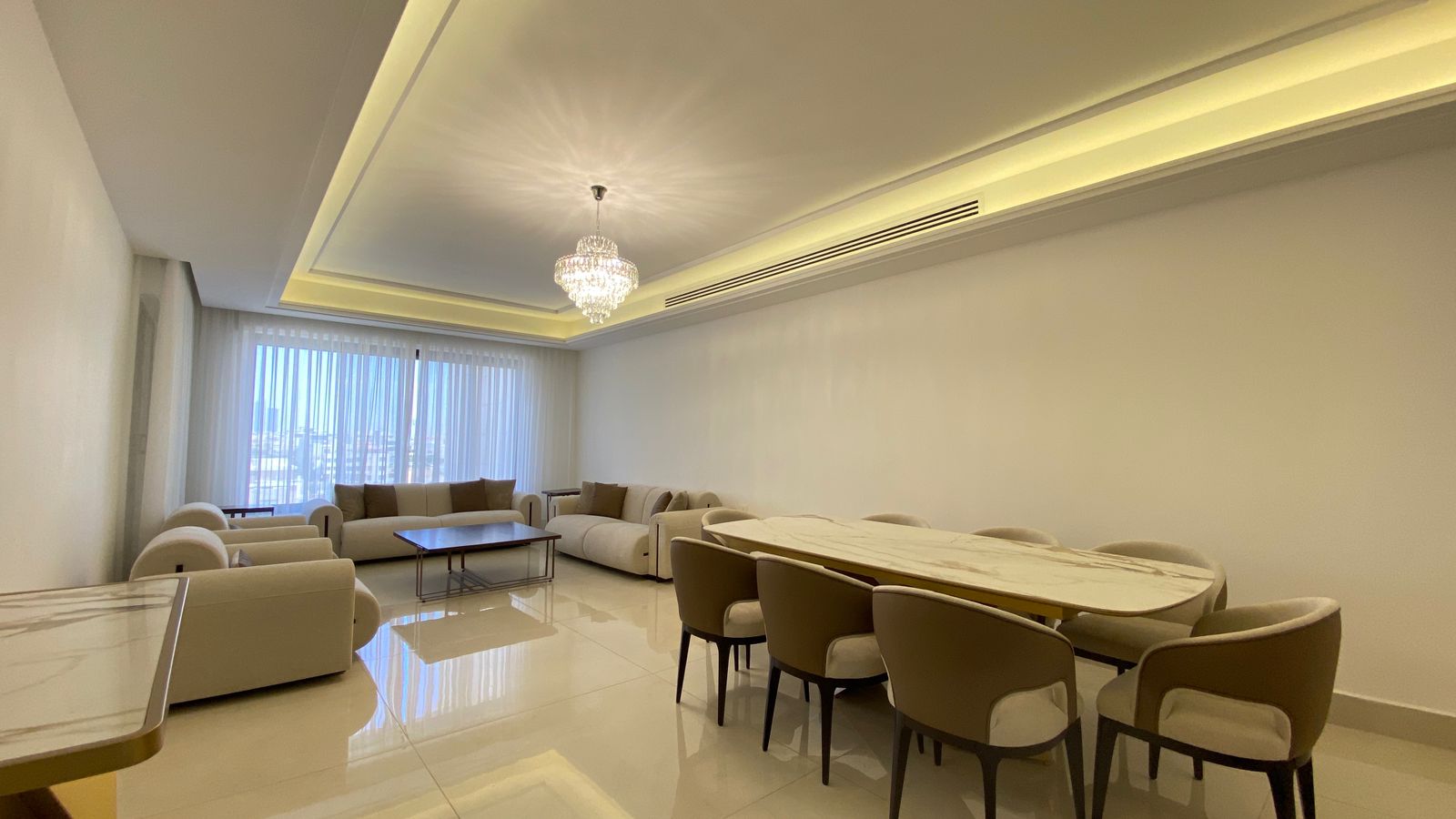 233da08b-c457-441f-aff8-f3398ff04f58 Spacious modern living and dining area with beige sofas, a marble dining table, and a crystal chandelier; large windows with sheer curtains illuminate the room.