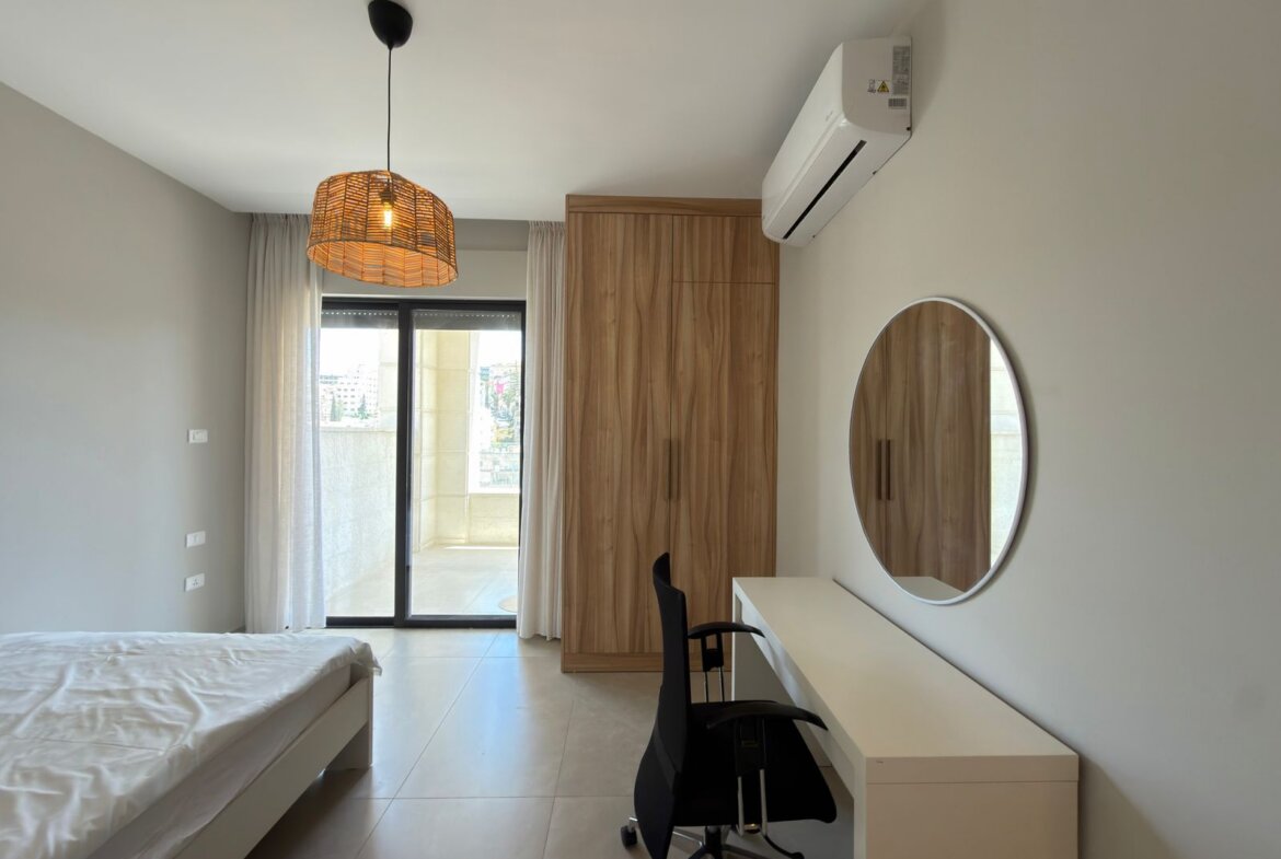 Modern bedroom with a bed, wooden wardrobe, white desk with black chair, circular mirror, and glass balcony door with sheer curtains; wall-mounted AC unit above.