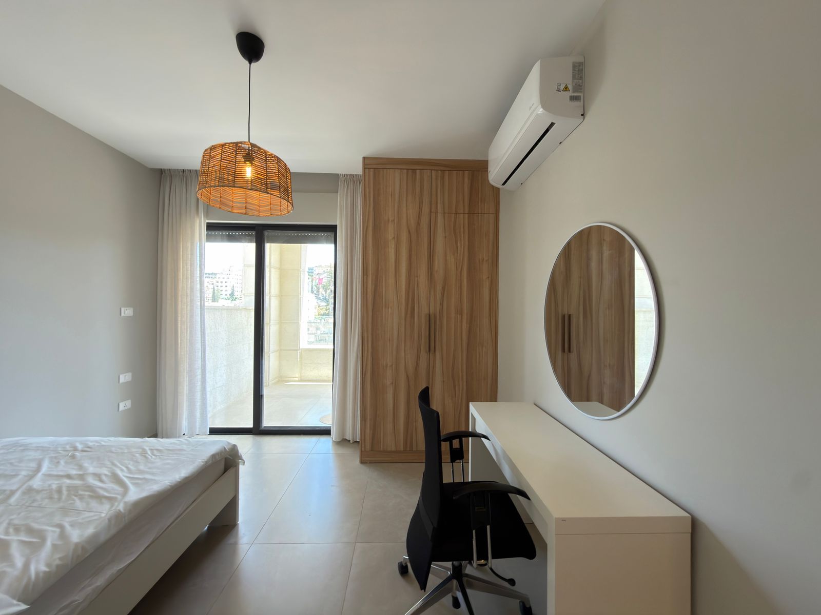 2360449e-2a07-40bf-b9c3-1774b0192a2c Modern bedroom with a bed, wooden wardrobe, white desk with black chair, circular mirror, and glass balcony door with sheer curtains; wall-mounted AC unit above.