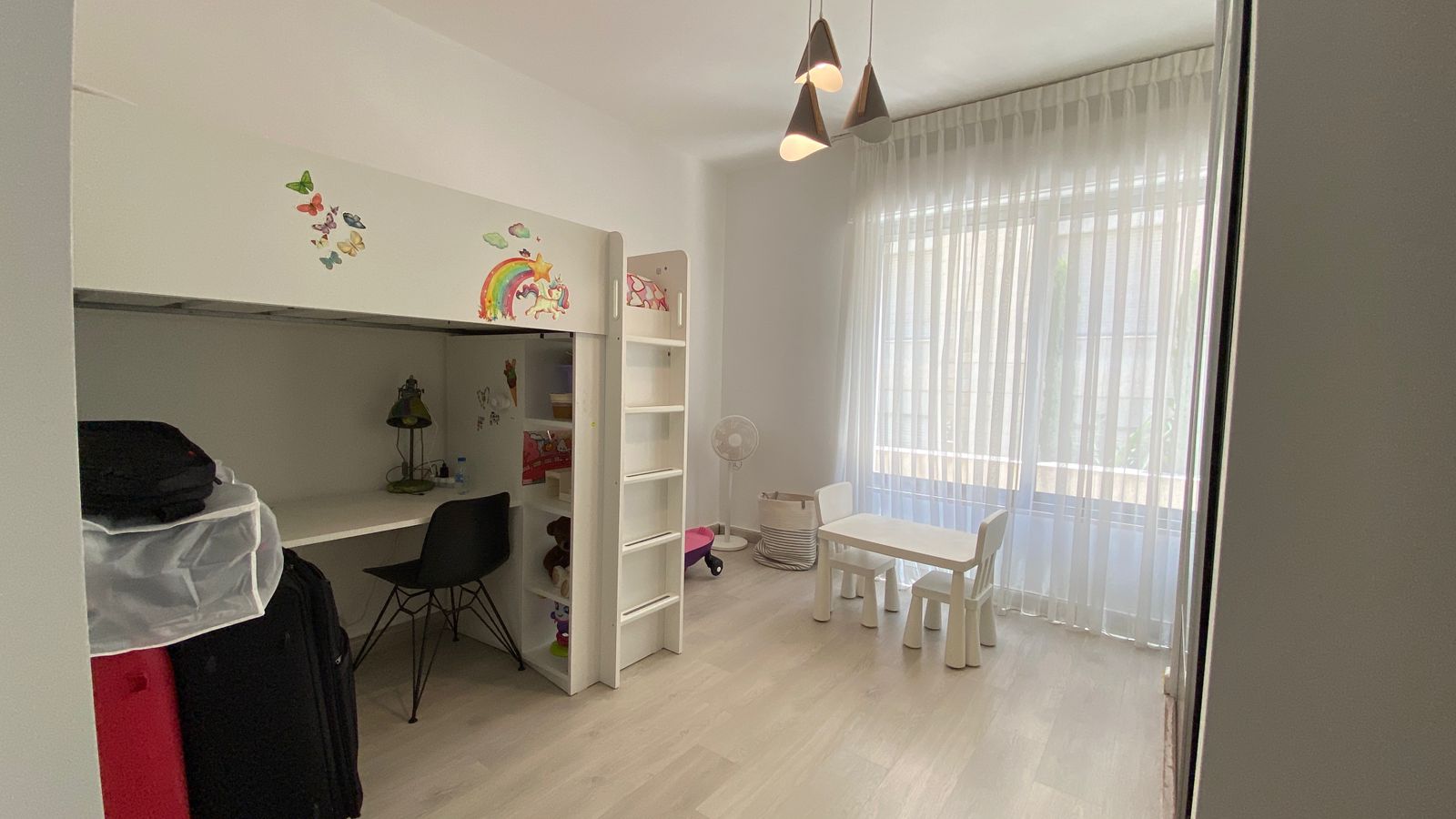 237108f2-48eb-4100-88bb-3fa6b15ed1d9 White loft bed with built-in desk in a bright kids' room, decorated with rainbow and butterfly wall decals, black desk chair nearby.