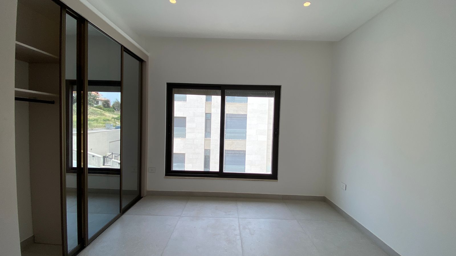 24b9647f-86bf-4544-ac67-f668f7c5abf2 Empty modern room with light tile floor, white walls, a large window, and a built-in sliding-door wardrobe on the left.